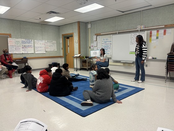 Students sit on the carpet while teachers stand with a book in front of them 