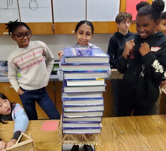 A group of students placing textbooks on their popsicle-stick bridge placed between two tables