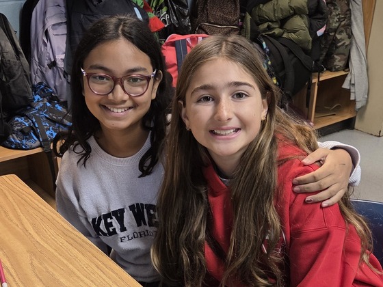 Two students posing at their desks. They are sharing a hug their big smiles show they are happy to be back in school.