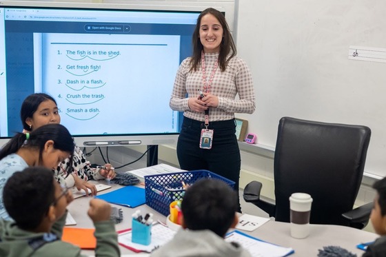 teacher stands in front of her class smiling while working on a lesson on the whiteboard