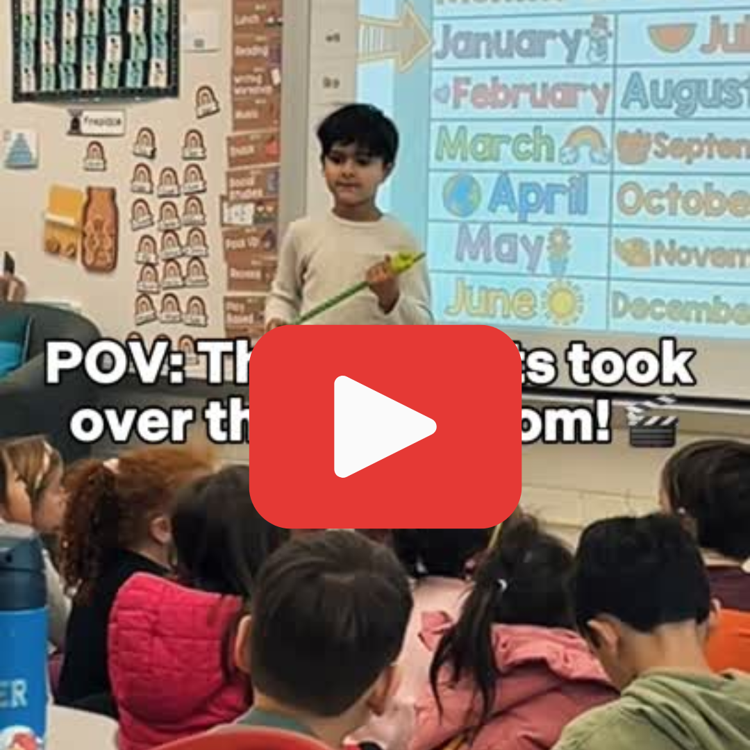 A student standing in front of a group of students who are sitting on the carpet. A red play button is overlayed.