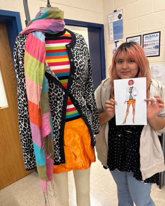 Student holding sketch if outfit design standing beside mannequin wearing design.