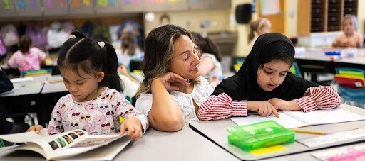a photo of a teacher in a classroom, helping two young students with their work.