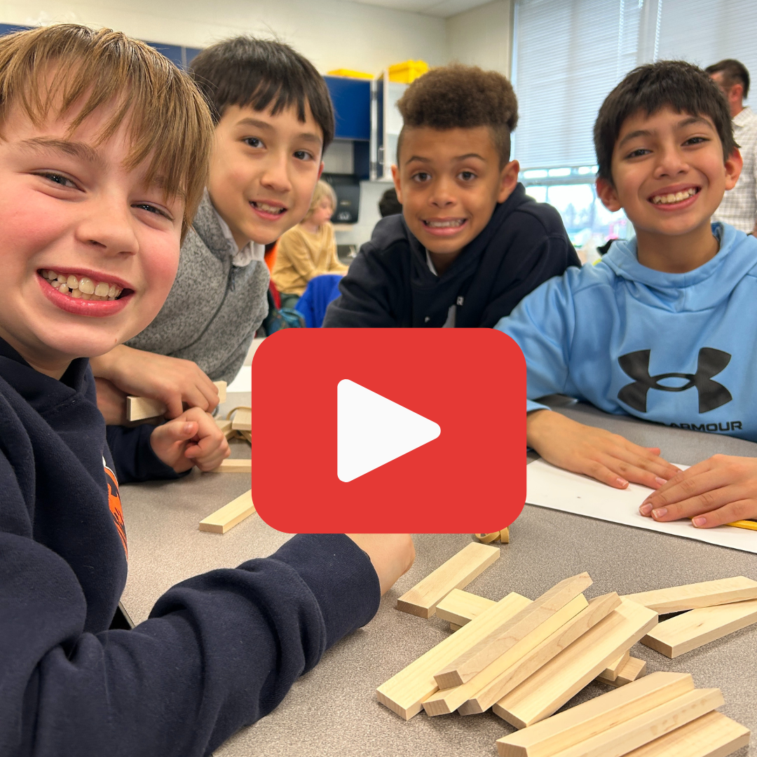 Four smiling elementary boys sit at a desk with wooden building blocks. A large red play button icon is centered over the image.