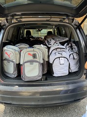 A car trunk sits open in a parking lot containing several rows of backpacks.