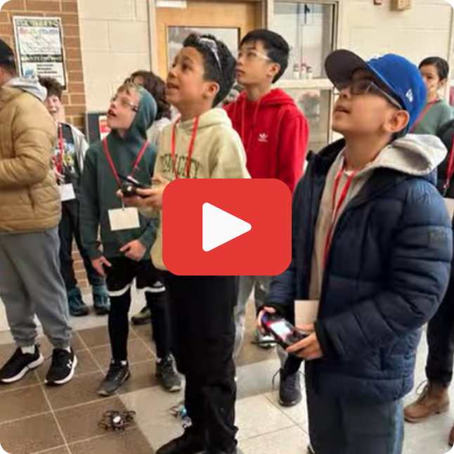 image of students looking up while holding controllers and flying drones inside school with red and white play button in the middle