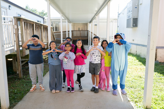 8  students (mix of boys and girls) under an overhang making the heart shape with their hands. Cheerful mood.