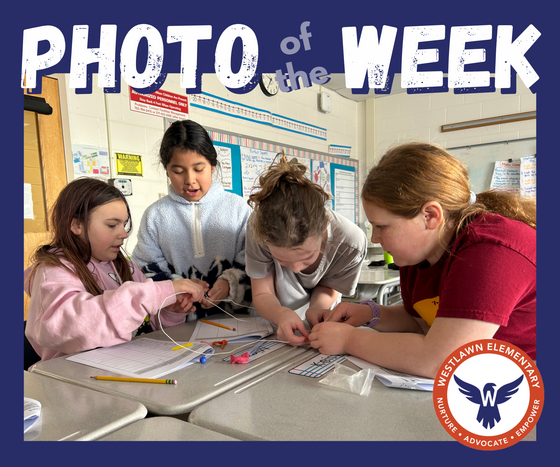 Photo of the Week: four students huddle together over an island made of desks and try to combine wires, a battery, and a lightbulb to create a circuit