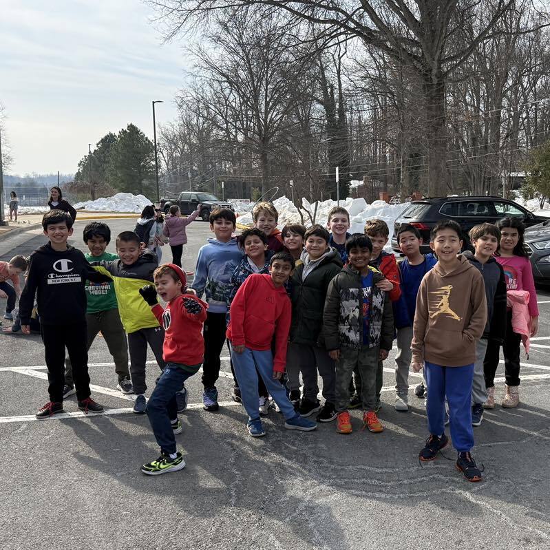 A large group of third-grade students pose together in a school parking lot on a sunny day with snow visible in the background.