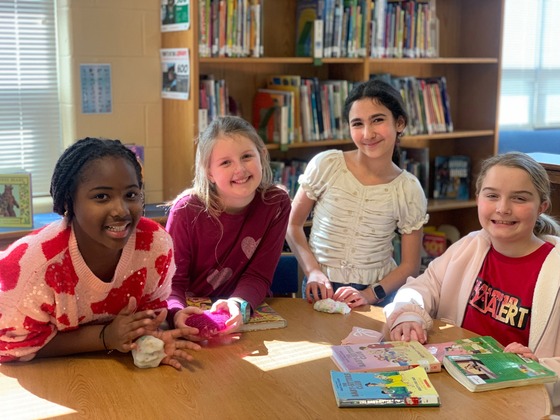 Four smiling girls gather at a sunny library table with Baby-Sitters Club books and small toys.