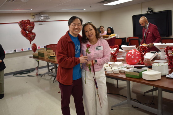 Dan Tra and a PTSA parent pose in front of the bagel and donut spread holding a rose