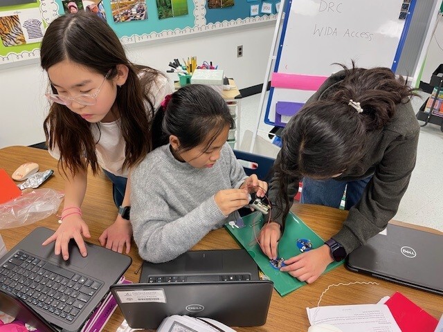 Three girls talking with each other with laptops in front of them on a table.