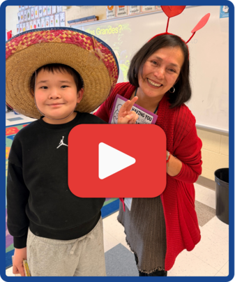 Video thumbnail of a student in a large straw hat and a teacher in red heart boppers smiling in a classroom.