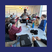 students sit at a table group, looking at their teacher who is soliciting suggestions from the class