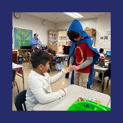 a student dressed like a superhero in a cape and mask hands a sticker to a first grader
