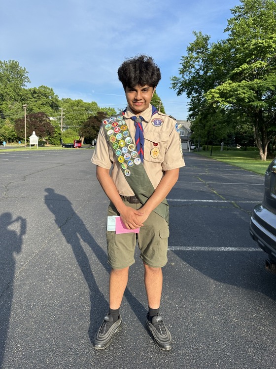 A teenage Scout in full uniform stands in a parking lot, wearing a sash filled with many colorful merit badges.