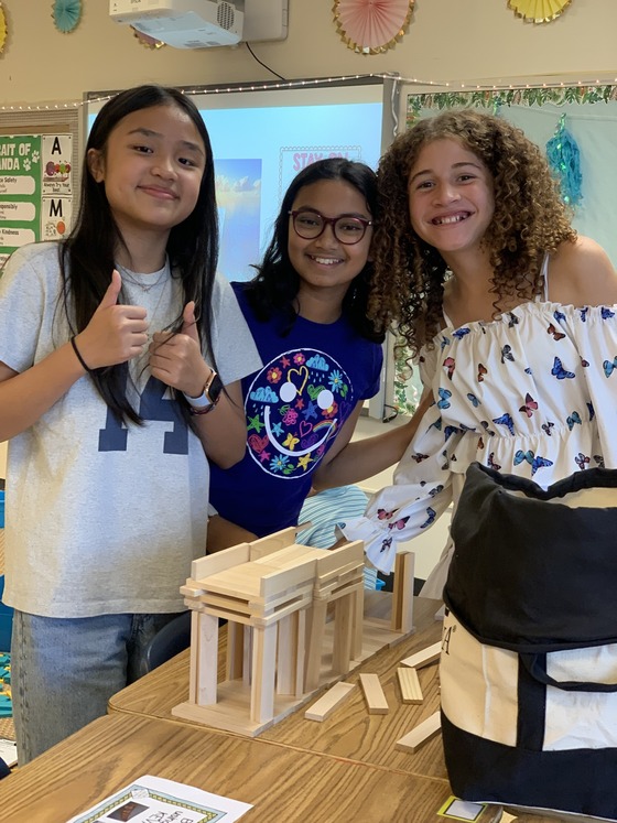 Three smiling students pose behind a tiered wooden block bridge structure they built on a classroom desk. One student gives two thumbs up.