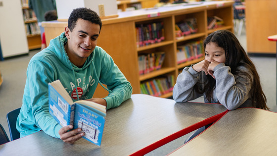 Mentor with an ES student at a table reading a book together.  Read the article below the image for more info.