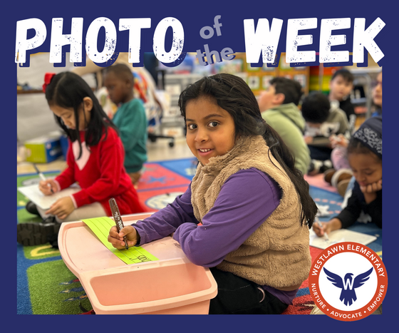 a first grade student smiles at the camera while she sits at a pink lap desk writing on a green sentence strip with a brown marker