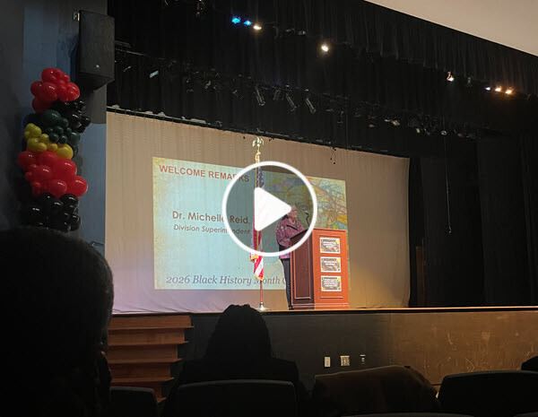 Superintendent on stage at a podium and there is a play button arrow on the image to see an embedded video