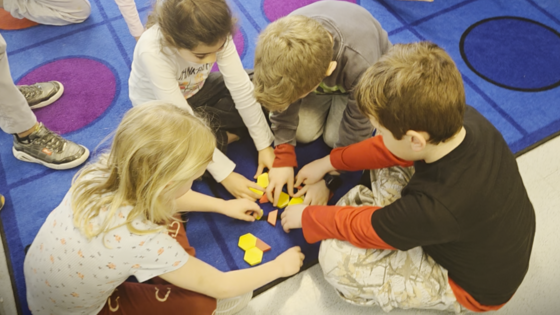 Four young students sit in a circle on a blue patterned rug, leaning in closely to work together on a shape puzzle.