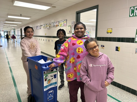 Four smiling elementary students pose together in a school hallway next to a large blue paper recycling bin.