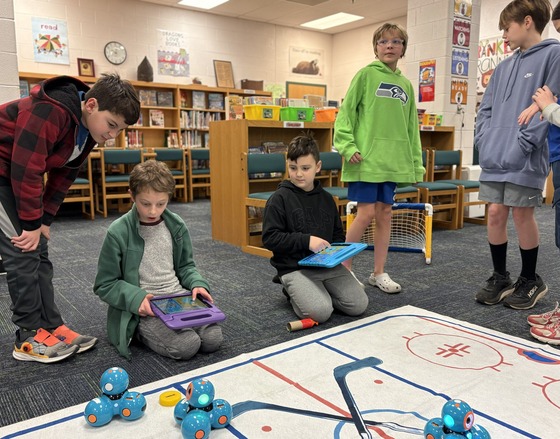 Five students using ipads to control small blue robots. 