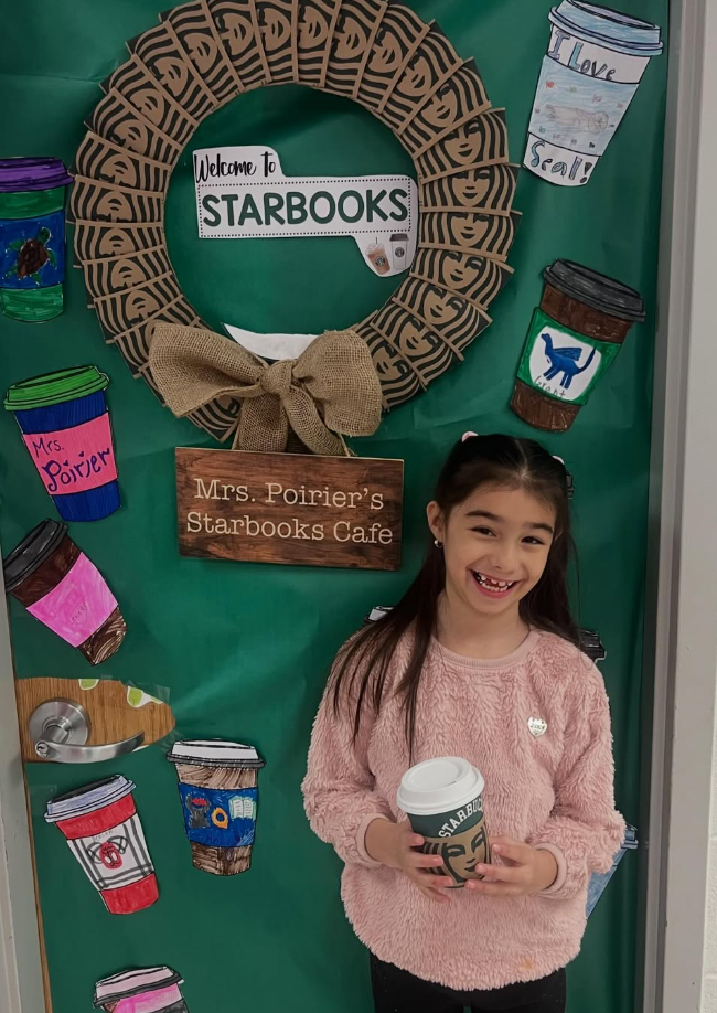 A smiling student holds a coffee cup in front of a green classroom door decorated with a "Starbooks" theme and colorful hand-drawn paper cups.