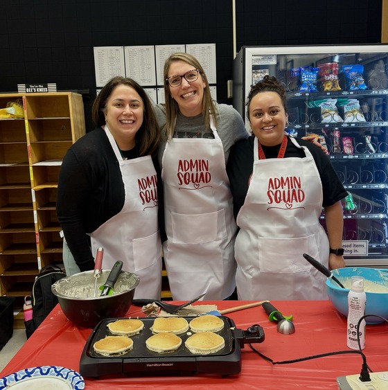 Photo of 3 female adults wearing white aprons that say Admin Squad with a hot plate of pancakes on the table in front of them.  