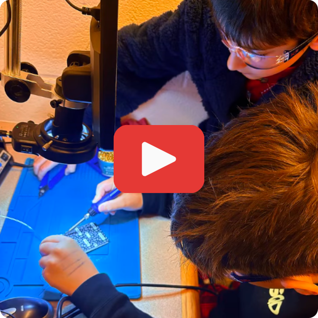 two boys at a soldering station working on an electronic device with a red and white play button on top of the image.