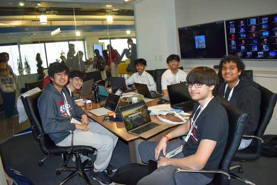 A group of TJ students sitting around a table in one of the HackTJ rooms, looking at camera and smiling.