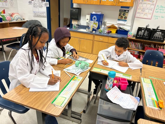 Three students in Young Scholars writing in notebooks while wearing white lab coats.