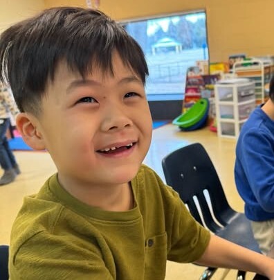 A smiling student looks up happily while coloring a picture at a classroom table.