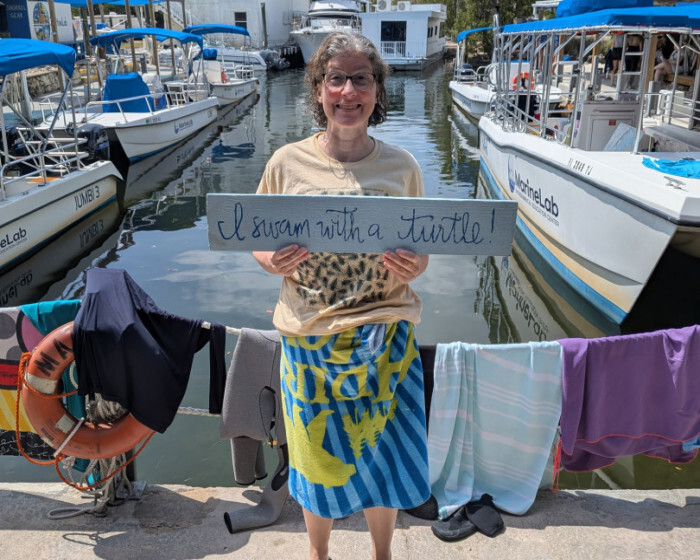 McLean HS Teacher, Julia Murdock poses on an ocean pier holding a sign which says "I swam with a sea turtle"