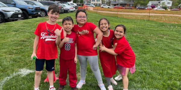 Five smiling children wearing red shirts pose together on a grassy field with cars and houses in the background.