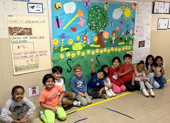 a picture of a row of students, sitting in a school hallway, underneath a sign that says "Spring Sprouts"