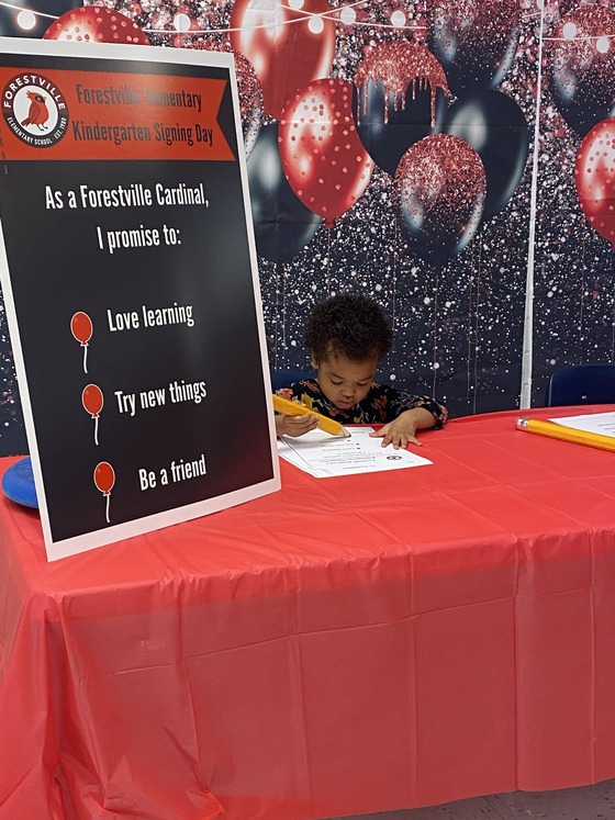 Young child signs a kindergarten pledge at a red table beside a sign about learning, trying new things, and being a friend.