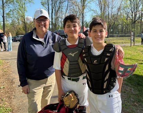 Adult and two students in baseball gear smile together on a field; one student wears catcher’s gear and holds a glove.