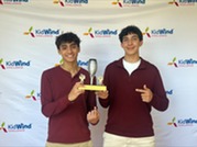 Two students pose with a trophy at a KidWind Challenge event; both point to the award.