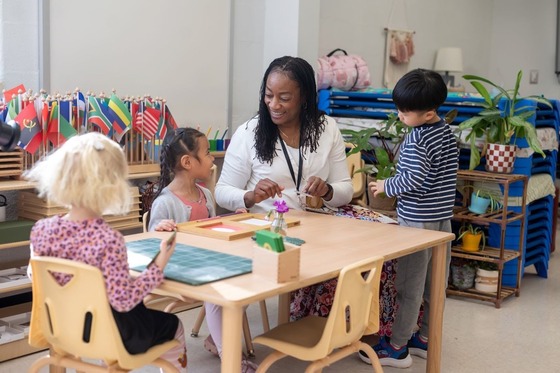 teacher sits at a small table with young students working on a craft
