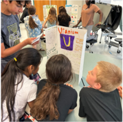 Four students huddled around a "Uranium" science fair board, curiously reading facts and looking at a hand-drawn atomic Bohr model.