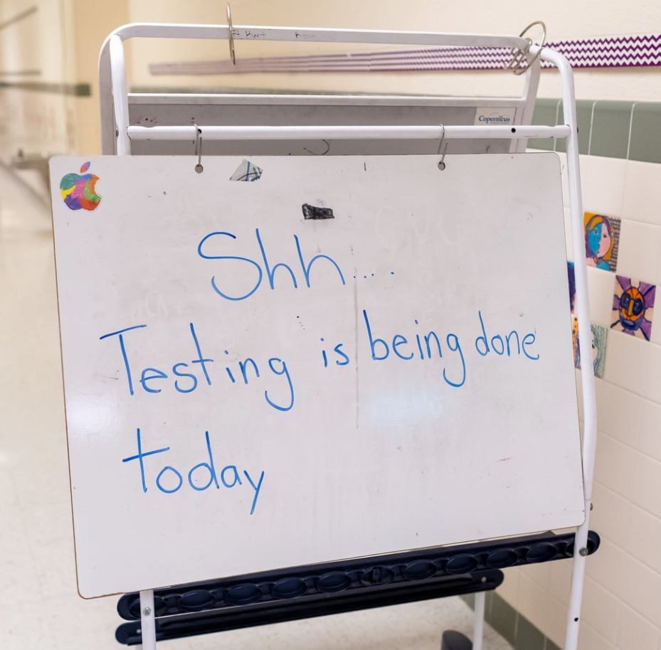 Whiteboard on a rolling stand in a school hallway reads, “Shh… Testing is being done today.”