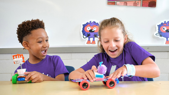 Two excited students in purple shirts build and play with colorful, custom-made toy rovers at a classroom table.