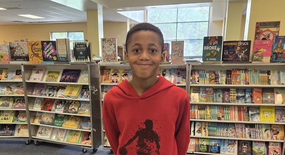 A smiling student wearing a red hoodie with a basketball graphic, standing in front of the book fair bookshelves set up in our library.