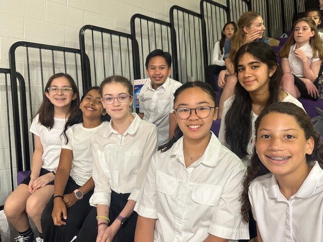 seven students smiling and sitting on bleachers in school gym with other students behind them