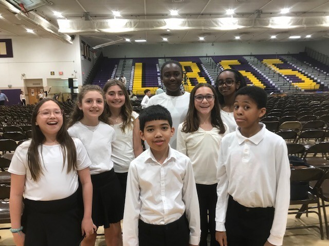 Seven 6th grade students in white shirts and black pants smile together in a gymnasium before a chorus festival.