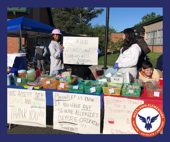 Students hold up posters at the bake sale table advertising the treats available for purchase