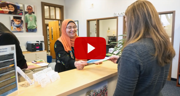 Screenshot of a vide featuring a woman behind a desk smiling and handing materials to another woman. 