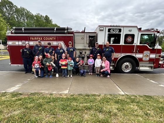 Preschool students and staff pose with firefighters in front of a Fairfax County fire truck, many children wearing firefighter helmets.