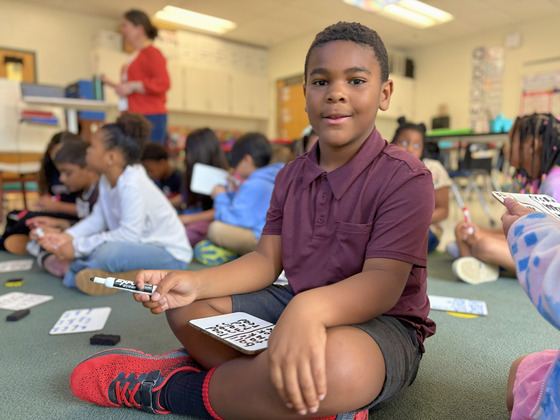 Student smiling in a classroom while practicing writing on a small handheld whiteboard.